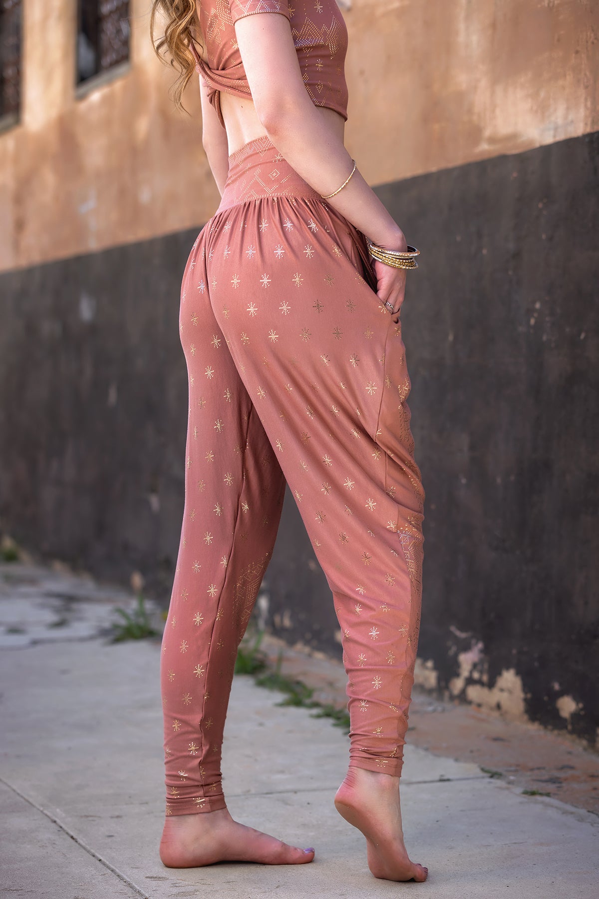 Barefoot on a sidewalk, a person wears Melodia Designs' Dendera Zaina Harem 2.0 Desert Rose Gold—relaxed-fit pants and matching cropped top in star-patterned pinkish fabric. Head not visible; textured wall in the background.