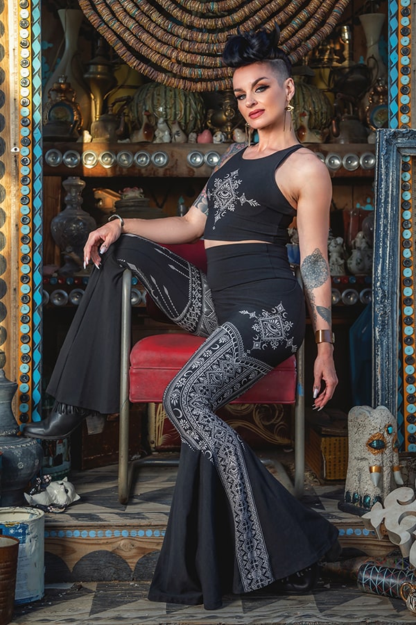 A woman with short dark hair poses confidently in an eclectic, vintage-decorated room, wearing Melodia Designs’ Tara Desert Dreams Pocket Flare leggings and a patterned crop top while seated on a red chair.