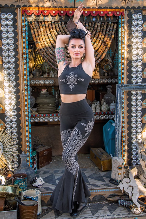 A woman wearing Melodia Designs Tara Desert Dreams Pocket Flare leggings and a black, mehndi-inspired crop top poses with arms raised in front of an ornate vintage shop filled with eclectic decorations.