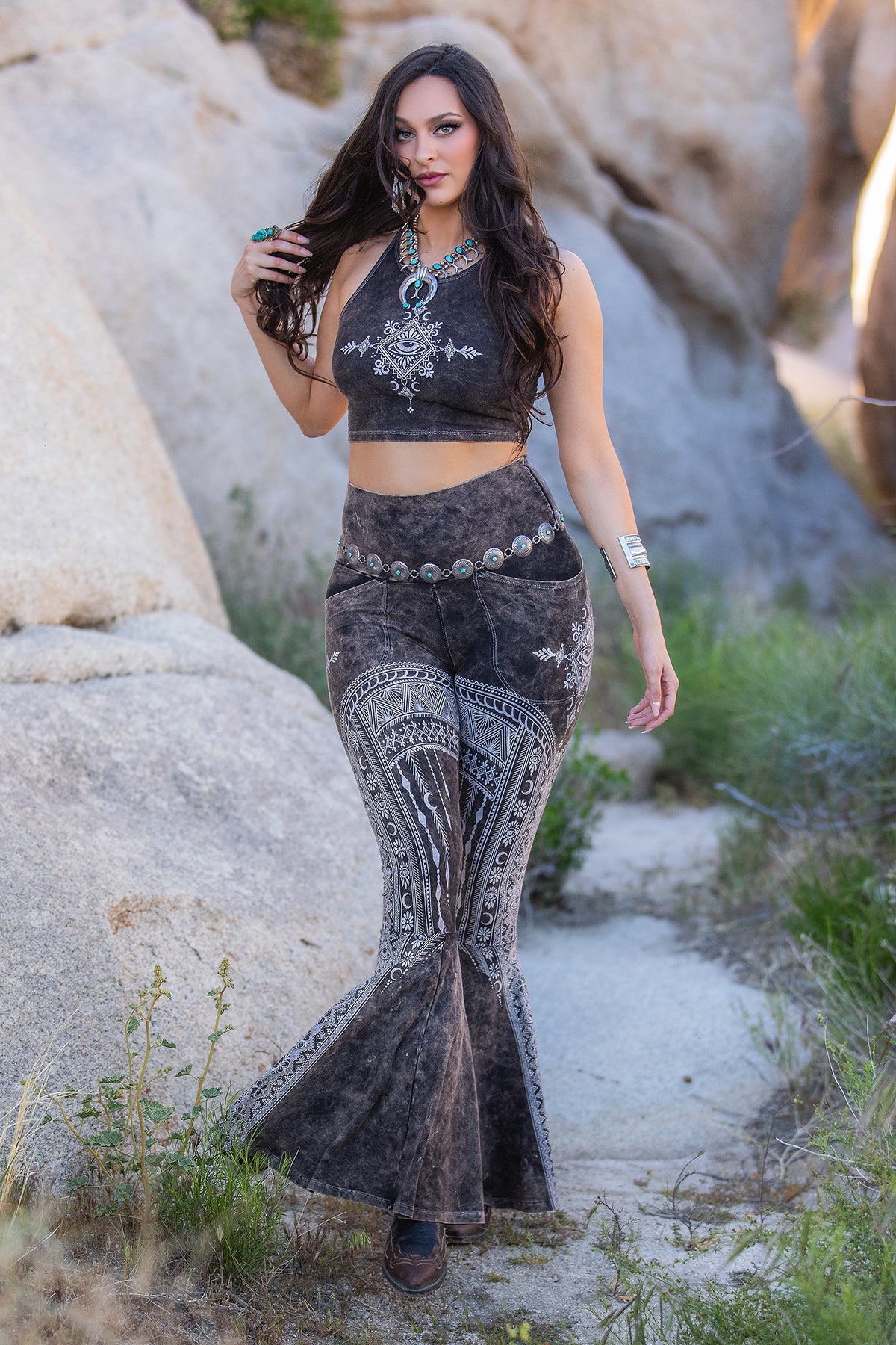 In a desert setting, a woman confidently walks among rocks and greenery wearing Melodia Designs Tara DD Pocket Flare Smoky Quartz leggings, paired with a patterned crop top, jewelry, and a belt.