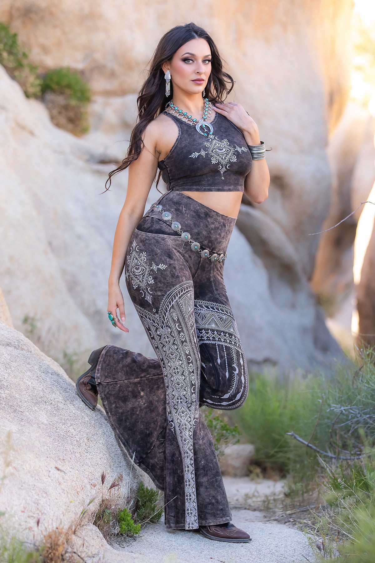 A woman poses on rocky terrain, wearing Melodia Designs Tara DD Pocket Flare Smoky Quartz set—eco-luxe fabric leggings with intricate white patterns and a matching boho crop top—accessorized with jewelry and sandals.