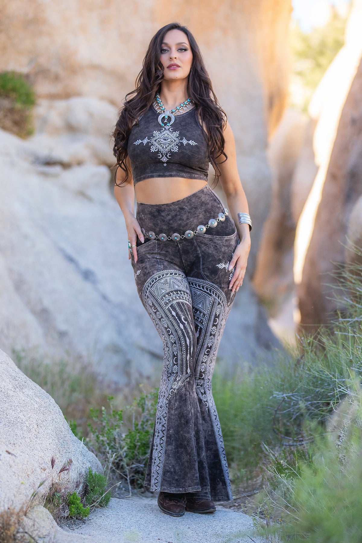 A woman stands among large rocks, wearing the Melodia Designs Tara DD Pocket Flare Smoky Quartz—sleeveless crop top and high-waisted flare leggings in eco-luxe fabric—styled with layered necklaces, bracelets, and a belt.