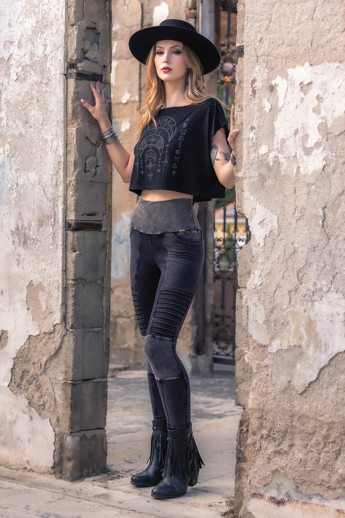 A woman in a black hat, Desert Oracle Dede Crop Top by Melodia Designs, and fitted pants with fringe boots stands confidently in a doorway framed by a weathered, textured wall, gazing at the camera. Her outfit is crafted from eco-friendly modal jersey, blending sustainability with effortless style.
