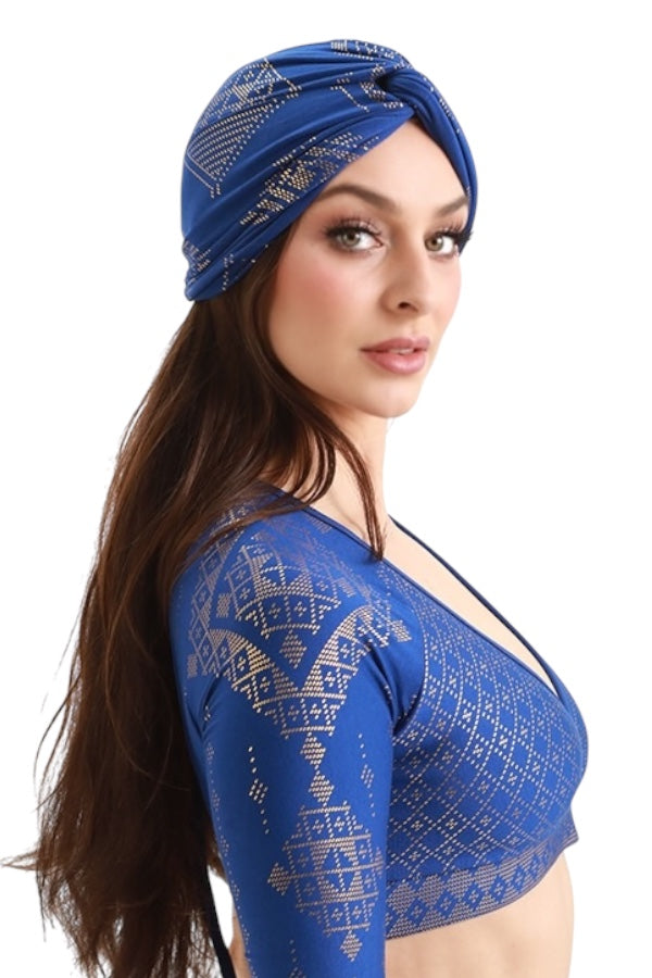 A woman with long brown hair models the Melodia Designs Urban Turban and a blue patterned wrap top, highlighting the styling versatility of this look as she glances over her shoulder against a white background.