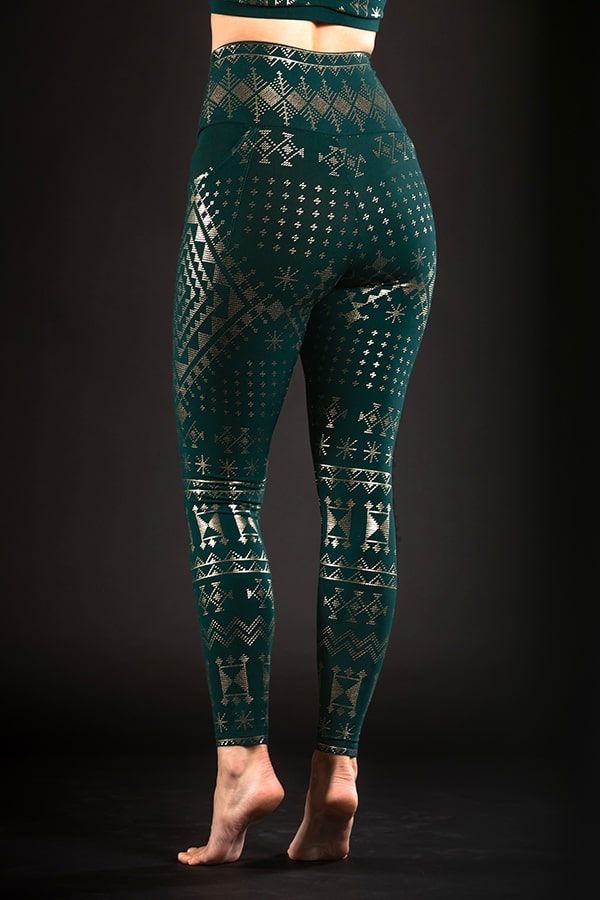 A person stands barefoot on tiptoes, showcasing the back of their legs in Melodia Designs Alkimiya Elsa Pocket Legging Malachite—green full-length leggings featuring gold geometric and snowflake patterns, crafted from eco-luxe fabric.