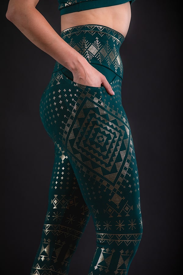 A person models Melodia Designs’ Alkimiya Elsa Pocket Legging Malachite, featuring gold geometric and star patterns, a high-rise foldable waistband, and side pockets; only their lower torso and upper thigh are shown against a black background.