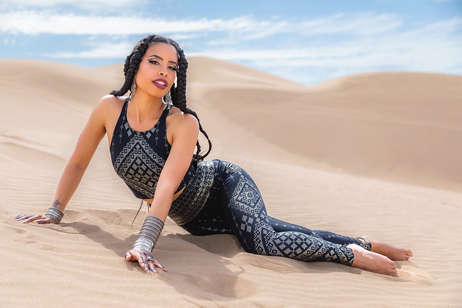 A woman with braids poses gracefully on sandy desert dunes in Melodia Designs’ Elsa Pocket Legging, featuring a black and gray pattern, under a blue sky dotted with clouds.