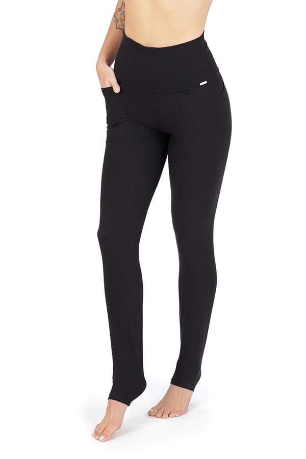 A person stands barefoot with one hand in the pocket of high-waisted black Elsa Pocket Leggings by Melodia Designs, featuring a foldable waistband and eco-luxe fabric. A tattoo is visible on their upper left arm; image cropped above the shoulders.