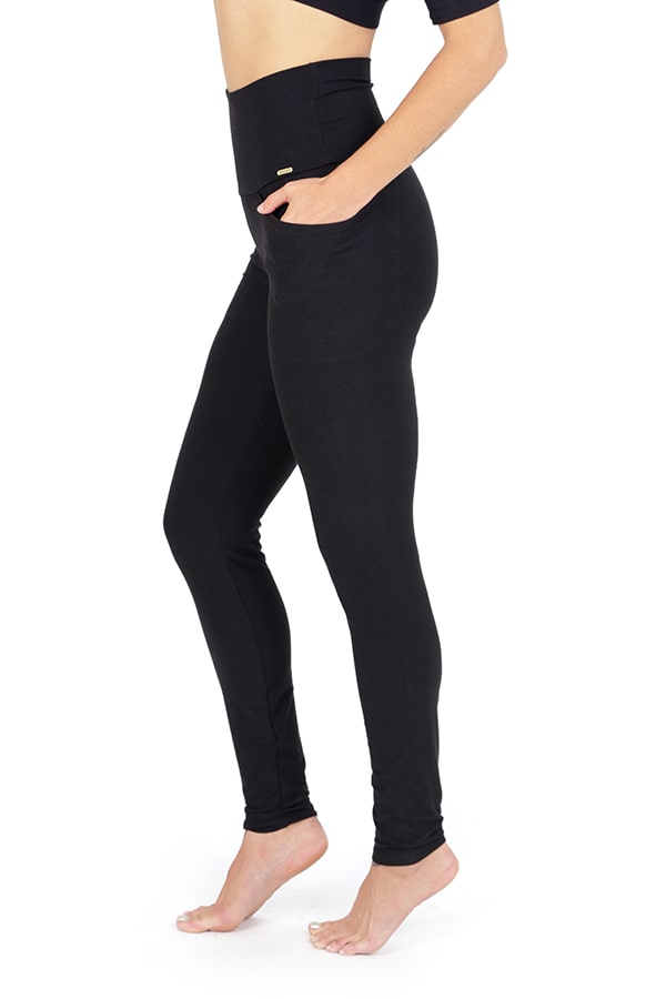 A person models Melodia Designs’ Elsa Pocket Legging—high-waisted black leggings with a foldable waistband and front pocket—paired with a black crop top, standing barefoot in profile against a white background.