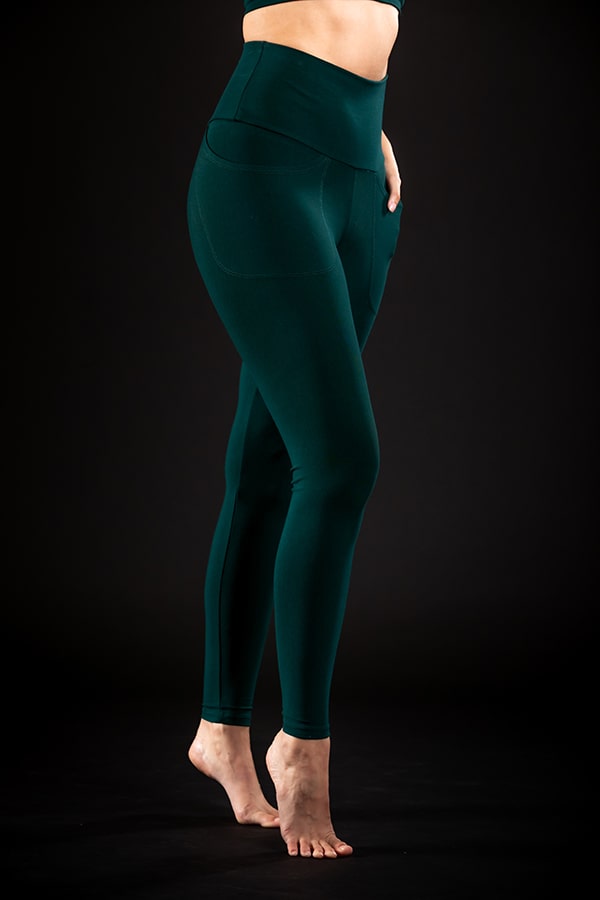 A person stands on tiptoes against a black background, wearing Melodia Designs Elsa Pocket Legging in Malachite—high-waisted, eco-luxe dark green leggings—with a matching sports bra. Their torso and head are not visible.