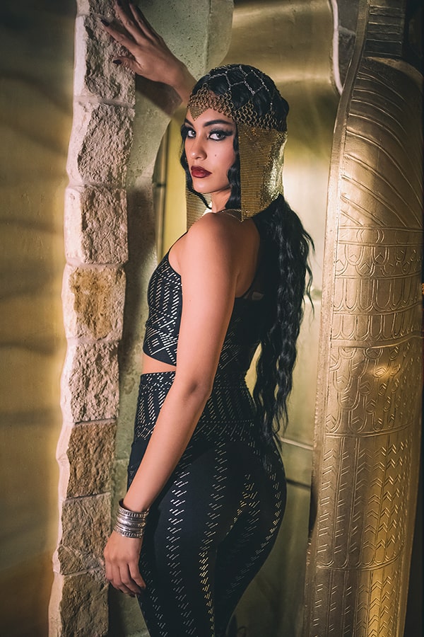 A woman wearing Melodia Designs Elsa 7/8 Pocket Legging PS and a gold headdress poses by a stone wall and golden sarcophagus, channeling an ancient Egyptian vibe.