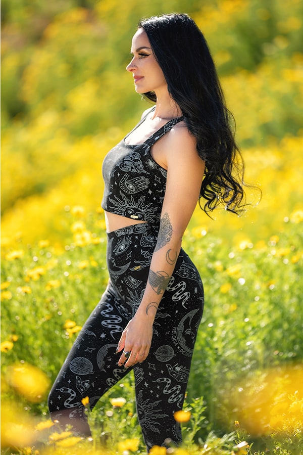 A woman with long black hair, wearing a black crop top and Melodia Designs Elsa Pocket Legging Celestial made from eco-luxe fabric, stands in a field of yellow wildflowers, gazing off to the side.