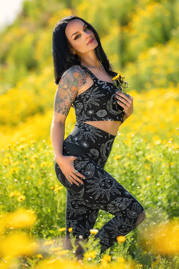 A woman with dark hair and tattoos poses confidently in a field of yellow flowers, wearing Melodia Designs Elsa Pocket Legging Celestial and a matching black patterned sports bra, holding small yellow flowers and looking at the camera.