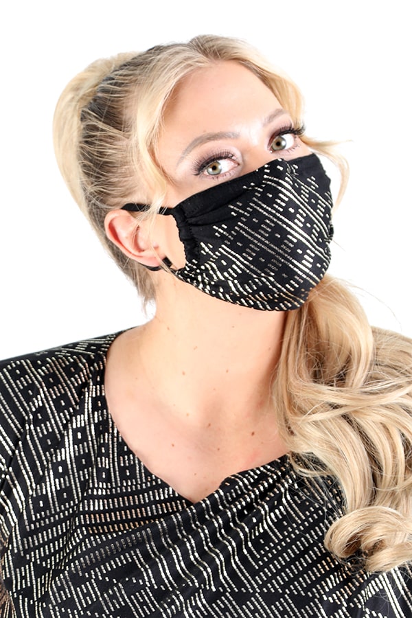 A woman with long blonde hair in a ponytail wears the black and gold patterned Festival Mask HM1 by Melodia Designs with a matching top, looking slightly upward. The background is white.