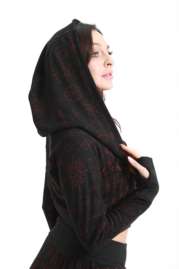 A person stands in profile against a white background, wearing the Melodia Designs Freydis Hoodie Runes— a black cropped zip-up hoodie with red bind rune-inspired geometric patterns— holding the hood and looking slightly upward.