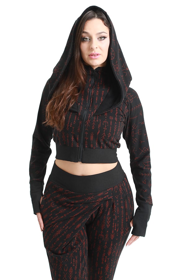 A woman stands with hands in pockets and hood up, wearing the Melodia Designs Freydis Hoodie Runes— a black cropped zip-up hoodie featuring red bind rune-inspired geometric patterns, paired with matching high-waisted pants.