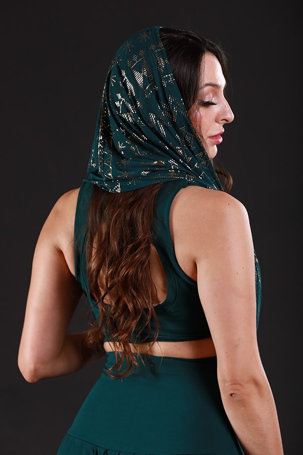 A woman with long wavy brown hair models the Melodia Designs Alkimiya Infinity Scarf Malachite, featuring gold patterns that shimmer over her head, paired with a dark green sleeveless outfit against a dark background.