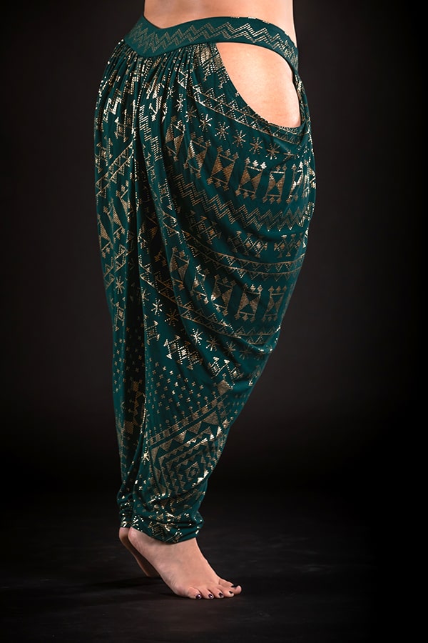 A person stands barefoot in Melodia Designs Alkimiya Isha Harem Pant Malachite—eco-luxe bamboo pants with green and gold patterns and large hip cutouts—against a black background.