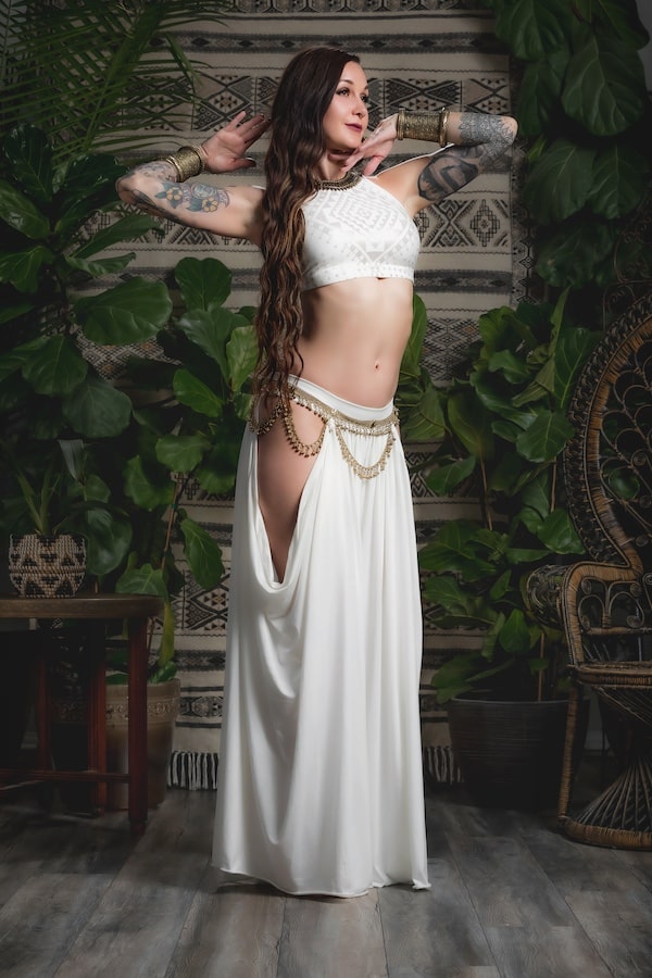 A woman poses indoors among lush plants and patterned decor, wearing Melodia Designs Aurora Peekaboo Palazzo Ivory, featuring a flattering waistband and graceful dance-inspired style.