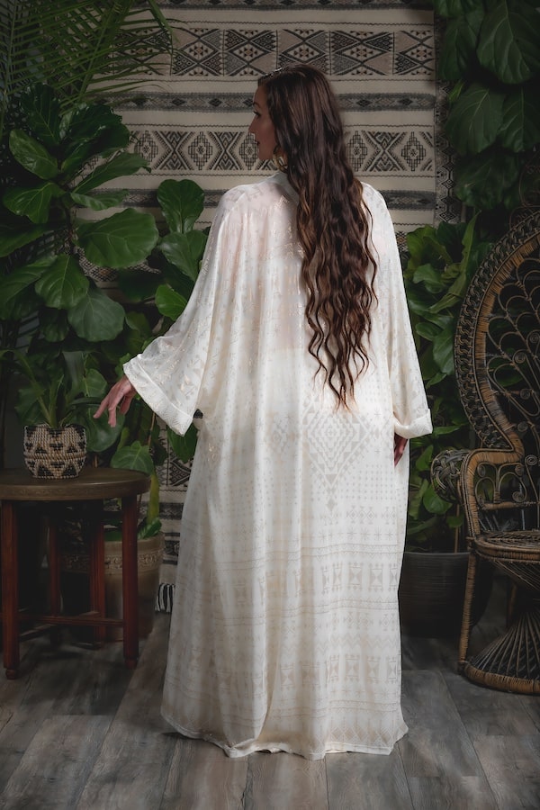 A woman with long wavy hair stands with her back to the camera, wearing the Alkimiya Zsa Zsa Robe Ivory by Melodia Designs. She is surrounded by lush green plants, woven furniture, and a decorative textile on a wooden floor.