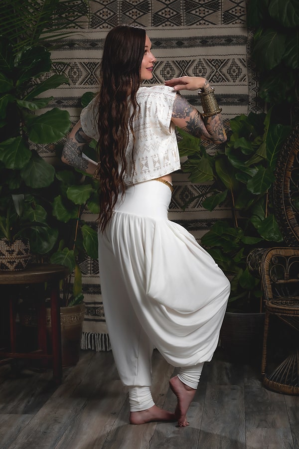 A woman with long brown hair and tattoos poses barefoot indoors in Melodia Designs Jazmine Harem Pant Ivory, paired with a white cropped top. She stands on one leg, arms raised, amidst lush green plants and patterned decor.