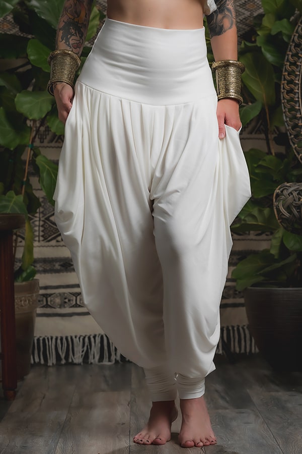 A person stands barefoot on a wooden floor wearing Melodia Designs Jazmine Harem Pant in Ivory, featuring a fold-over waistband and deep side pockets. They have tattoos, gold cuffs, and are surrounded by green plants and patterned decor.