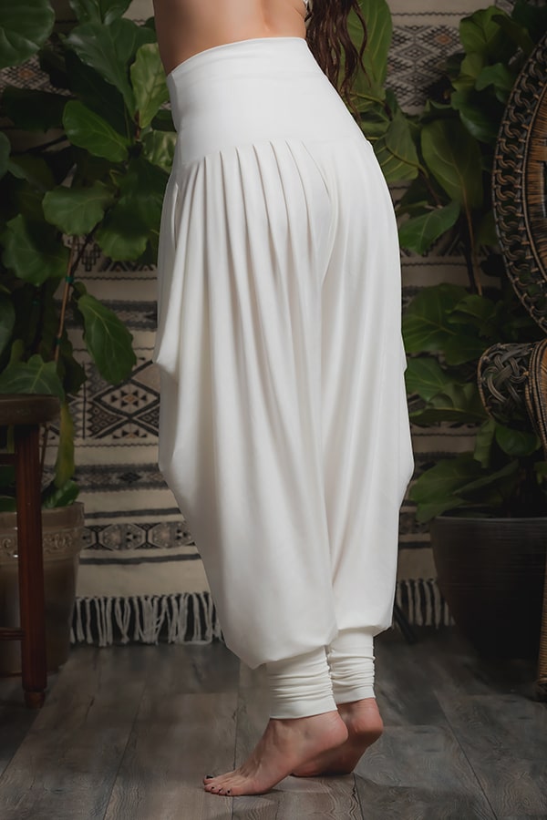 A person stands barefoot on a wooden floor, wearing Melodia Designs Jazmine Harem Pant in Ivory, featuring deep side pockets. Their upper body is out of frame, with green plants and patterned textiles in the background.