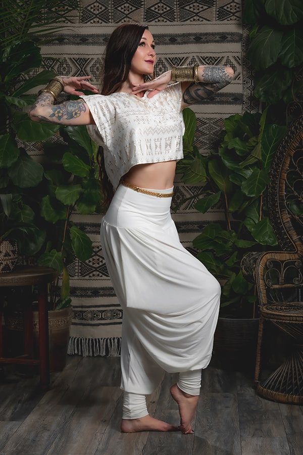 A woman with long hair and tattoos poses gracefully indoors in an eco-luxe white crop top and Melodia Designs Jazmine Harem Pant Ivory, standing on one foot among green plants and patterned textiles, arms bent in elegance.