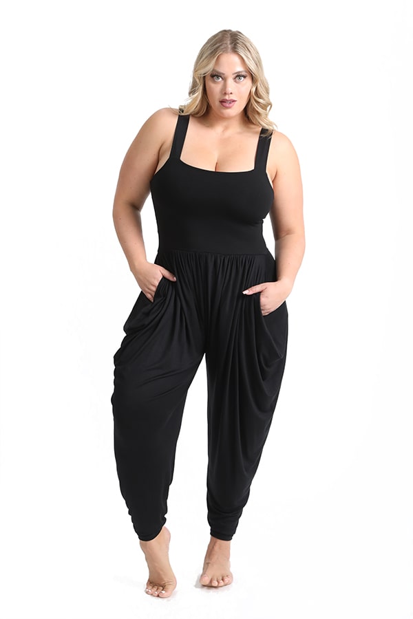 A woman with blonde hair models the Melodia Designs Jinni Jumper: a sleeveless black jumper with wide straps, harem pant–style loose-fit, and pockets. She stands barefoot against a plain white background, looking confidently at the camera.