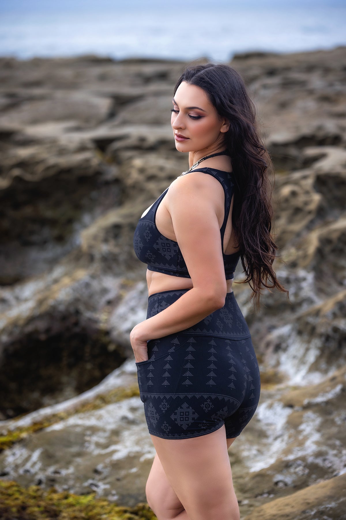 A woman with long dark hair stands on rocky terrain, facing sideways. She wears a dark sports bra and Melodia Designs’ Karnak Alice Short Shadow, made from eco-luxe fabric with geometric patterns, looking over her shoulder calmly.