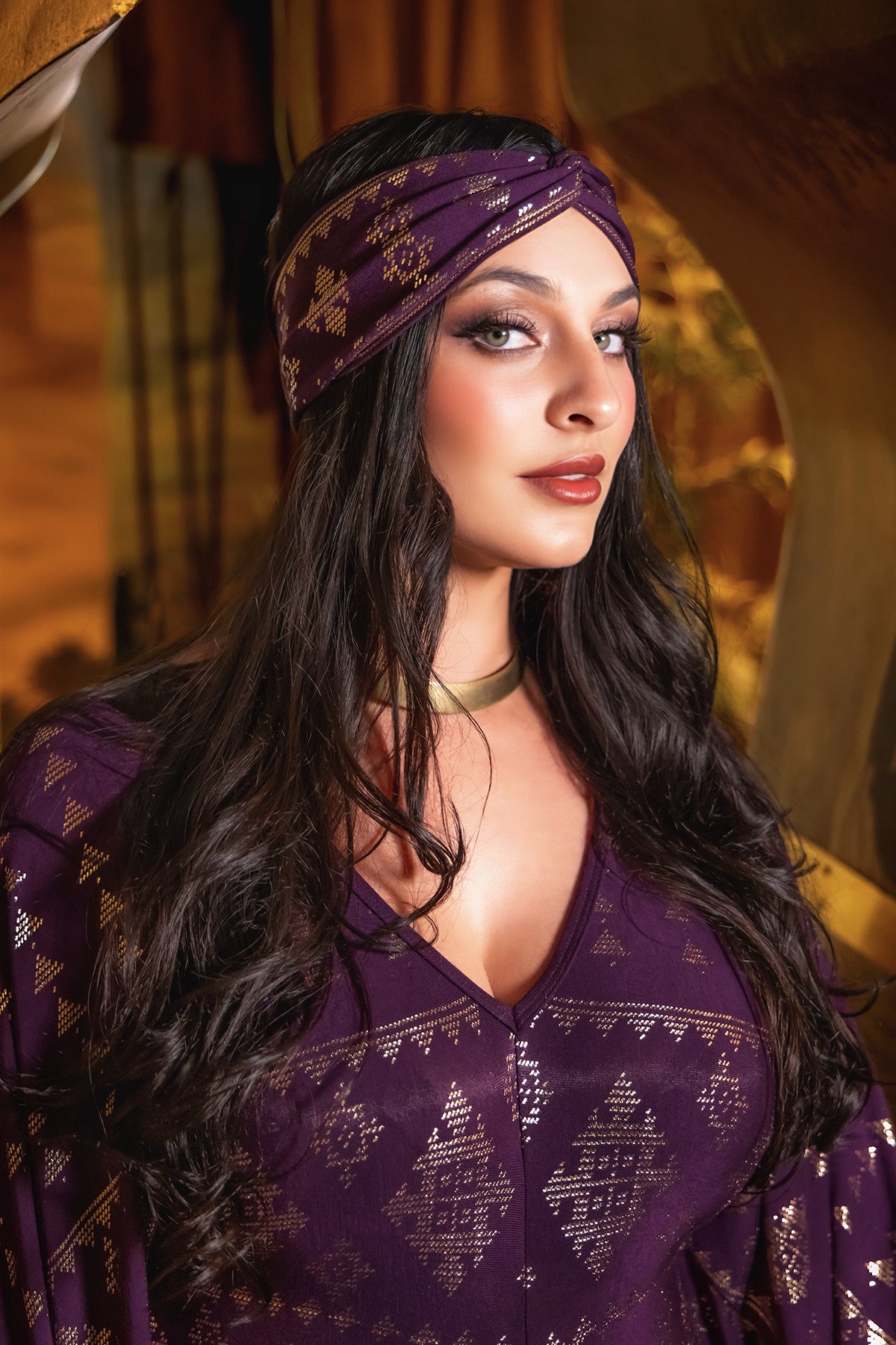 A woman with long dark hair poses confidently indoors, wearing the Melodia Designs Karnak Contessa Caftan 2.0 in Aubergine-Gold with a matching headband and gold accents, deep-V neckline, and a gold choker necklace. She gazes slightly to the side.