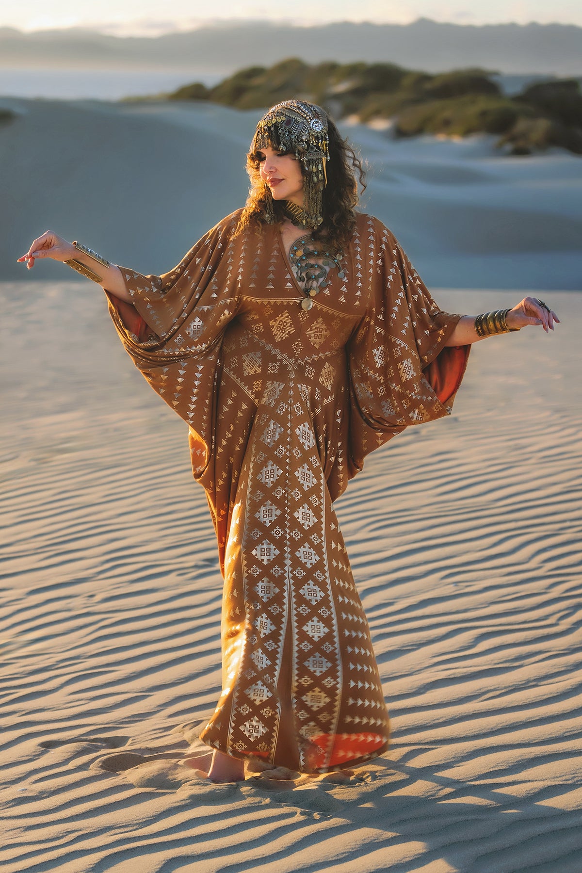 A woman barefoot on sand dunes wears the Melodia Designs Karnak Contessa Caftan 2.0 Copper, a relaxed brown and white dress with batwing sleeves, ornate jewelry, and raises her arms as the sun casts long shadows across the landscape.