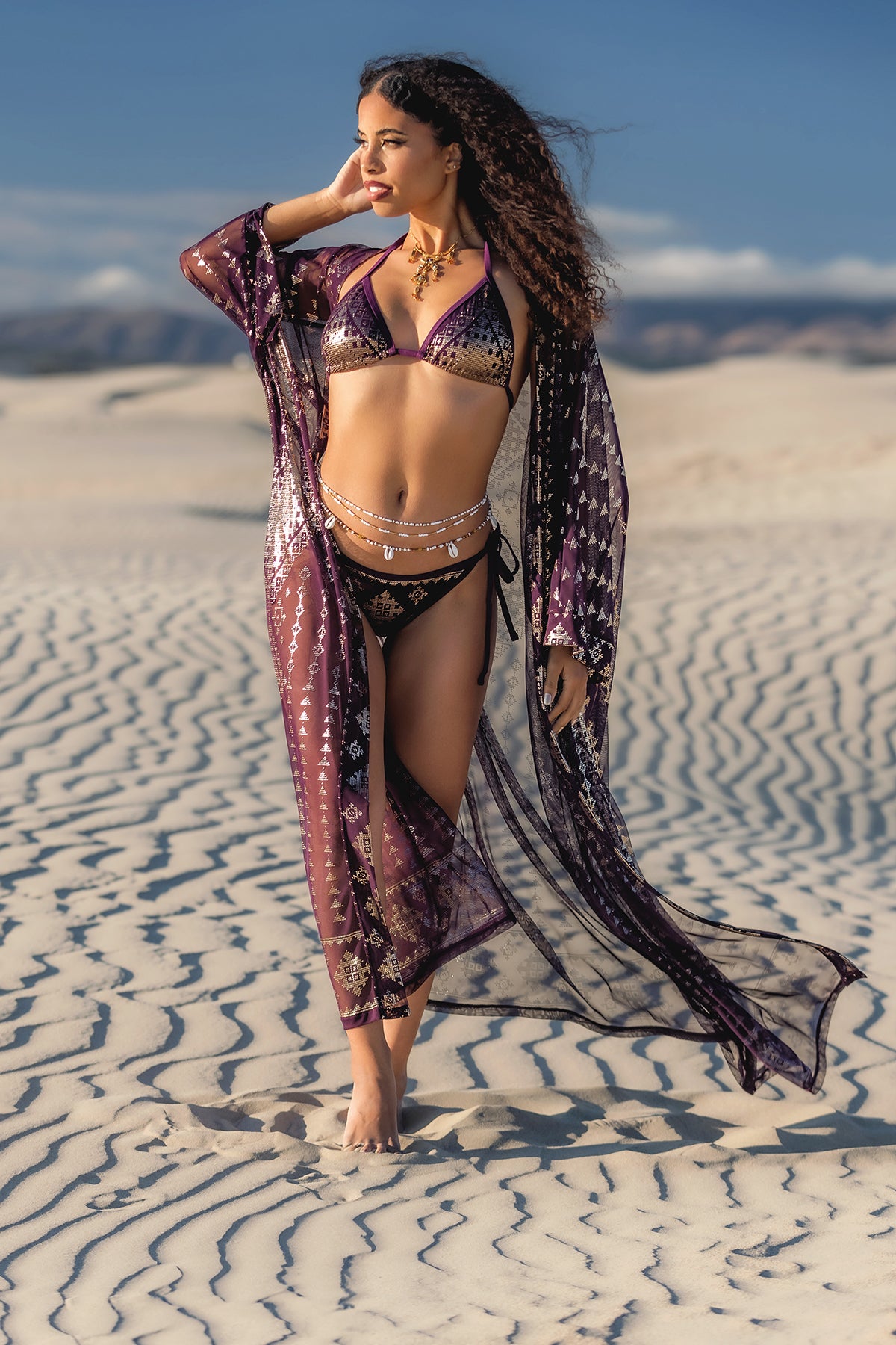 A woman in a Melodia Designs Karnak Zsa Zsa Aubergine-Gold robe with gold accents over a leopard print bikini stands barefoot on rippled sand, gazing to the side and touching her hair under the bright sun.