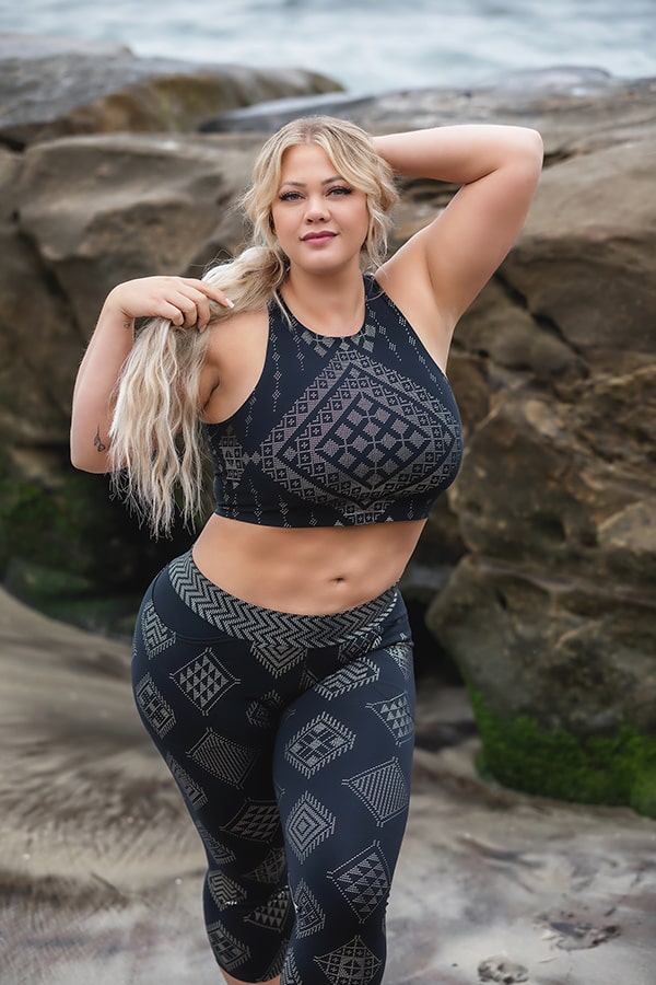 A woman with long blonde braided hair poses on rocky seaside terrain wearing the Laura Crop by Melodia Designs and leggings featuring geometric patterns in high-performance fabric for a sporty look.