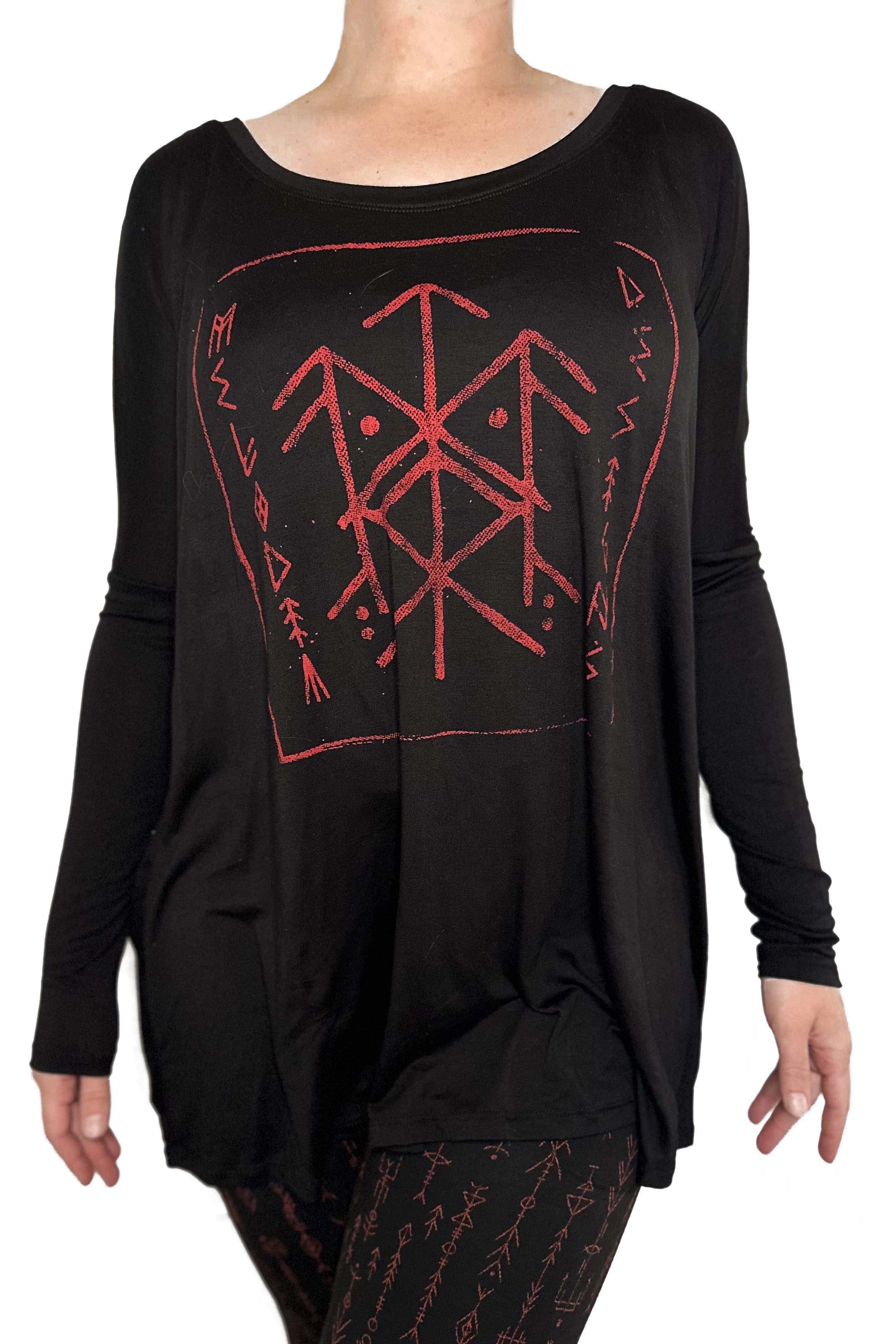 Someone wears the Melodia Designs Luna Long Sleeve Runes, a loose black top with red bindrune-inspired geometric patterns, paired with black rune-patterned leggings. Both are crafted from earth-friendly modal.