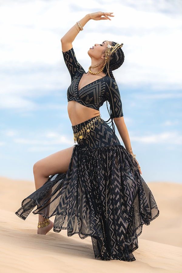 A woman poses gracefully on a sandy dune in the Golden Skirt Luxor by Melodia Designs—an elegant black and gold-patterned 1940s-inspired Egyptian dance skirt—her flowing skirt and gold jewelry gleaming under a blue sky with wispy clouds.