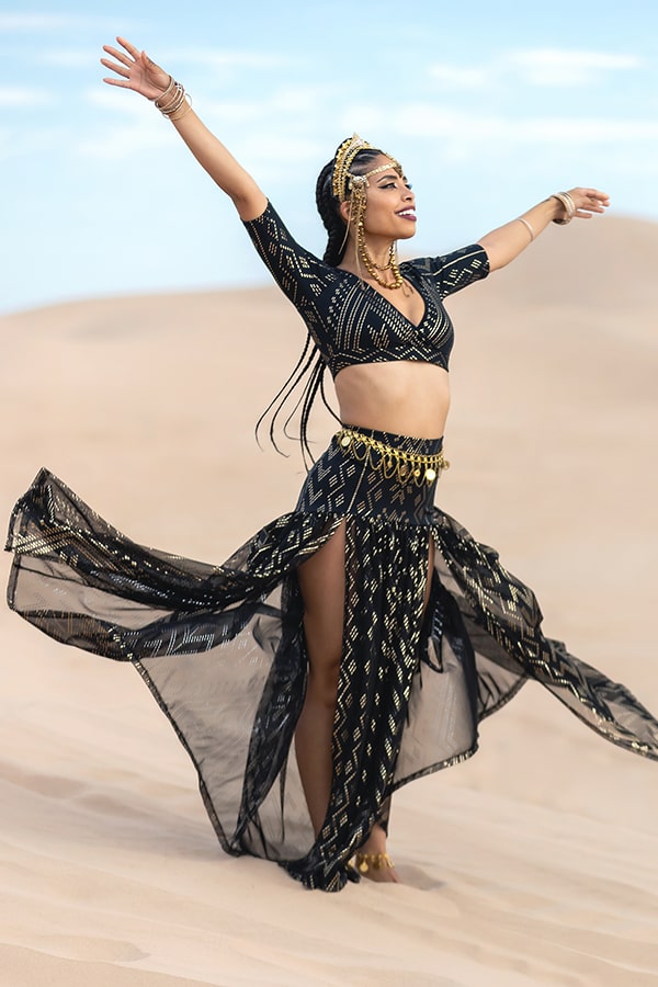 A woman stands barefoot on sand dunes, arms raised joyfully, wearing the Melodia Designs Golden Skirt Luxor with a metallic belt and headpiece, black and gold crop top, long braids, and gold jewelry against a vibrant blue sky.