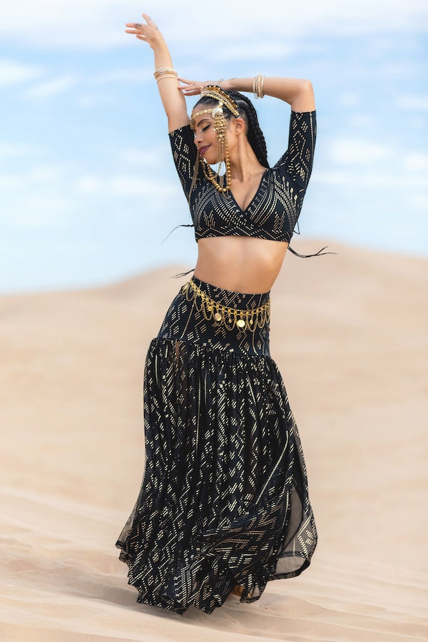 A woman in Melodia Designs Golden Skirt Luxor—a black and gold two-piece with a shimmering motif skirt—dances on sand dunes, adorned with gold jewelry and a headpiece, her arms raised beneath the blue sky.