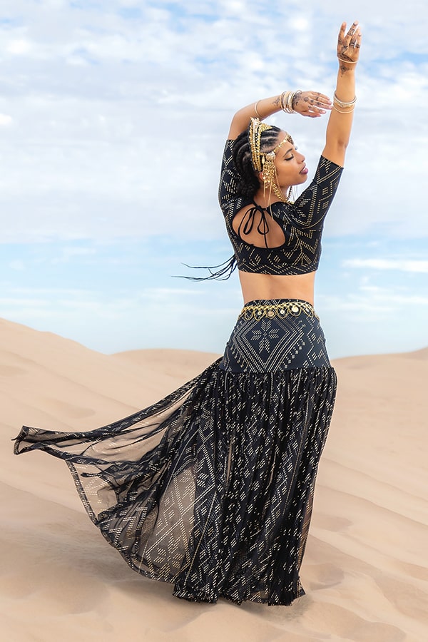 A woman stands on sand dunes with arms raised, her back to the camera, wearing Melodia Designs’ Golden Skirt Luxor—a black and gold shimmering motif skirt with a versatile waistband. The sky above is mostly clear with a few clouds.