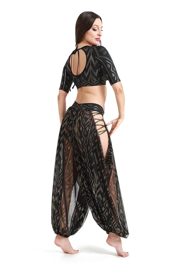 A woman poses barefoot in a sheer black geometric outfit featuring Melodia Designs’ Sahara Peekaboo Luxor Pantaloon with a draped hip, elastic-free waistband, and open-back crop top, set against a white background.