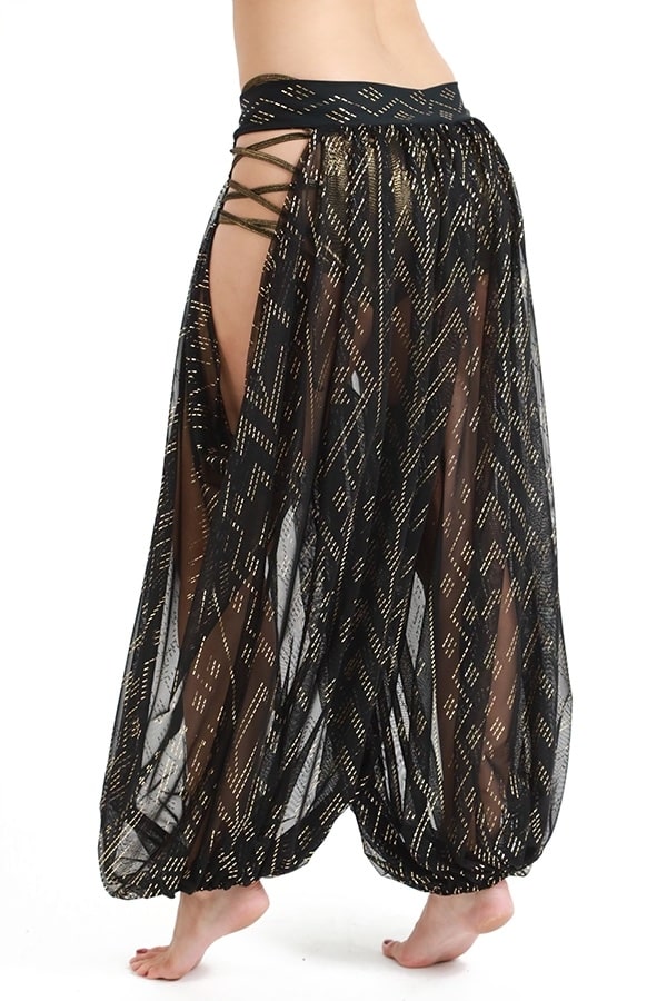 A person models Melodia Designs Sahara Peekaboo Luxor Pantaloon—loose, sheer black harem pants with gold geometric patterns, crisscross side slits, draped hip details, and an elastic-free waistband—standing barefoot against a white background.