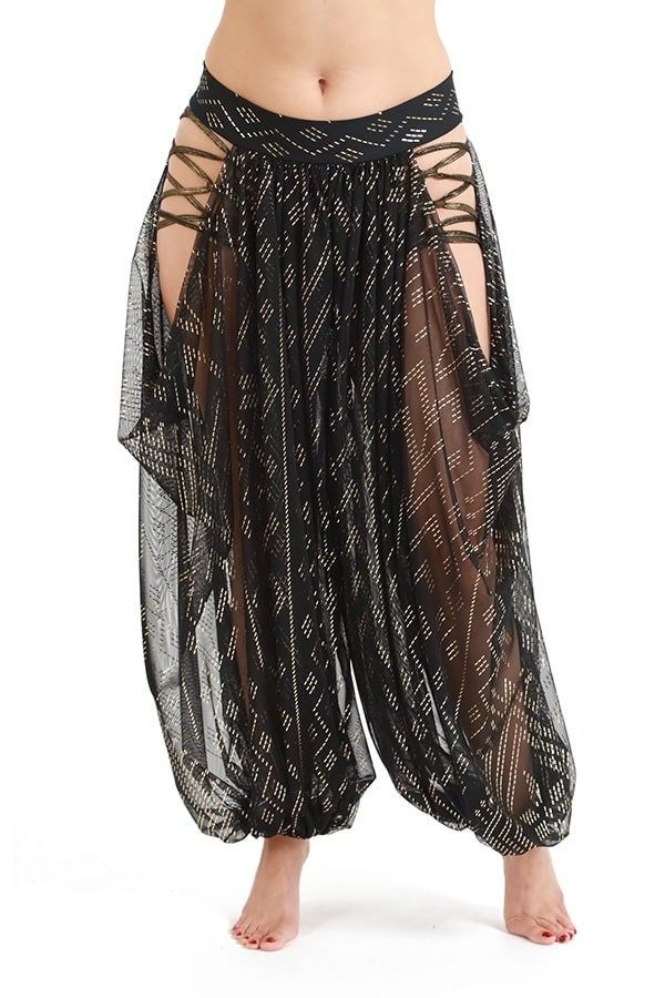 A person wearing Melodia Designs black Sahara Peekaboo Luxor Pantaloon with gold geometric patterns, high side cutouts, draped hip, and crisscross lace-up details stands barefoot against a white background.