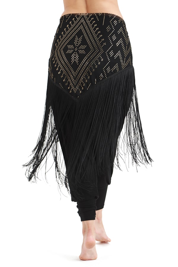 Back view of a person wearing the Melodia Designs Fringe Shimmy Shawl Luxor—black with gold-studded geometric patterns and long silky chainette fringe—making a bold statement barefoot against a white background.