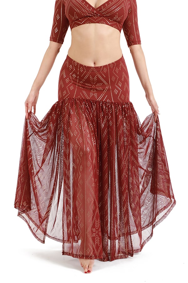 A person wears the Melodia Designs Golden Skirt Luxor, a maroon two-piece with a fitted crop top and flared skirt featuring a sheer geometric pattern, holding out the skirt against a plain white background.