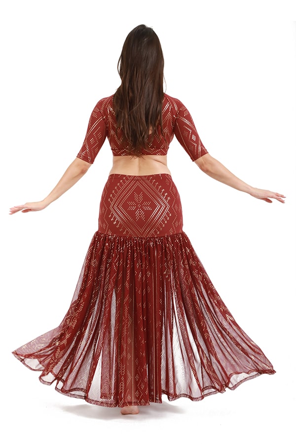 Woman with long brown hair stands barefoot with arms outstretched, wearing the Melodia Designs Golden Skirt Luxor, facing away from the camera against a white background.