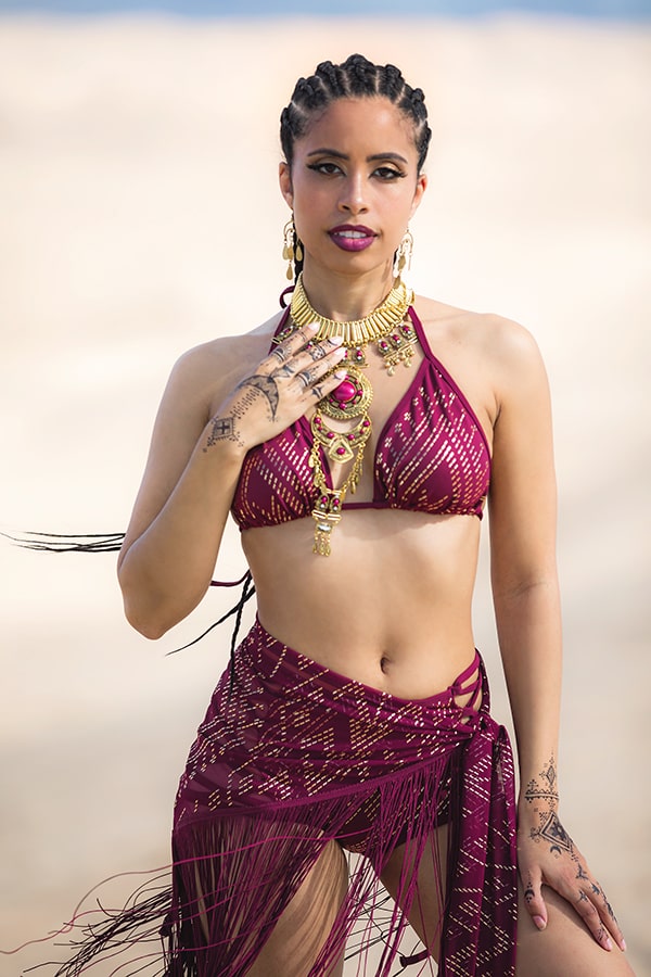 On a sandy beach, a woman in Melodia Designs Fringe Shimmy Shawl Luxor and a burgundy bikini shines—gold jewelry and braid hairstyles adding flair. The silky chainette fringe makes this shawl a standout festival fashion statement.