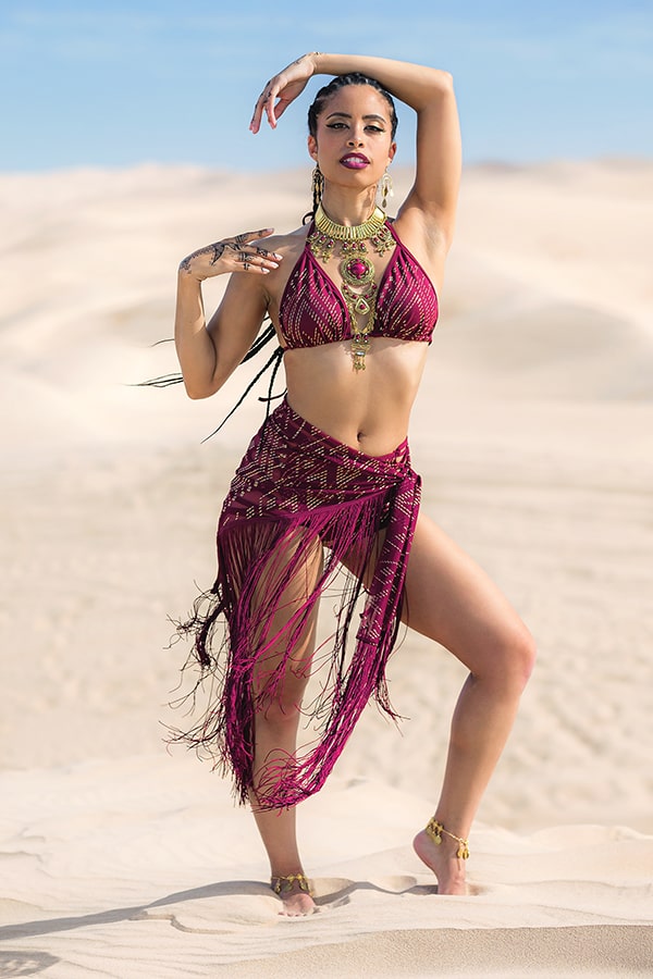 A woman stands barefoot on desert sand, one arm raised, wearing Melodia Designs Fringe Shimmy Shawl Luxor—magenta with fringe—paired with gold jewelry and anklets. The festival-ready look is set against a clear sky backdrop.