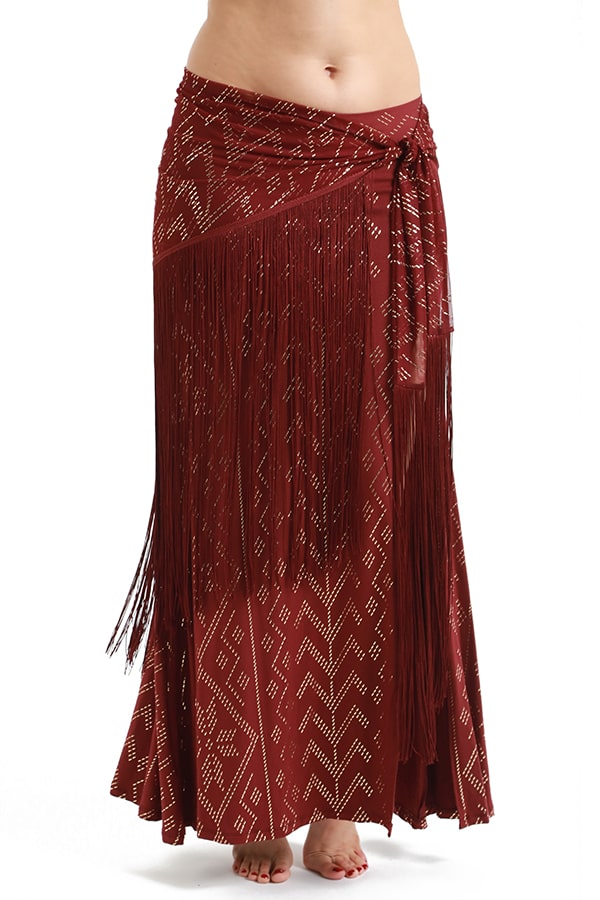 A person showcases the Melodia Designs Fringe Shimmy Shawl Luxor—a maroon piece with white geometric patterns and chainette fringe, tied at the waist and reaching the ankles, making a bold festival statement with bare feet visible.