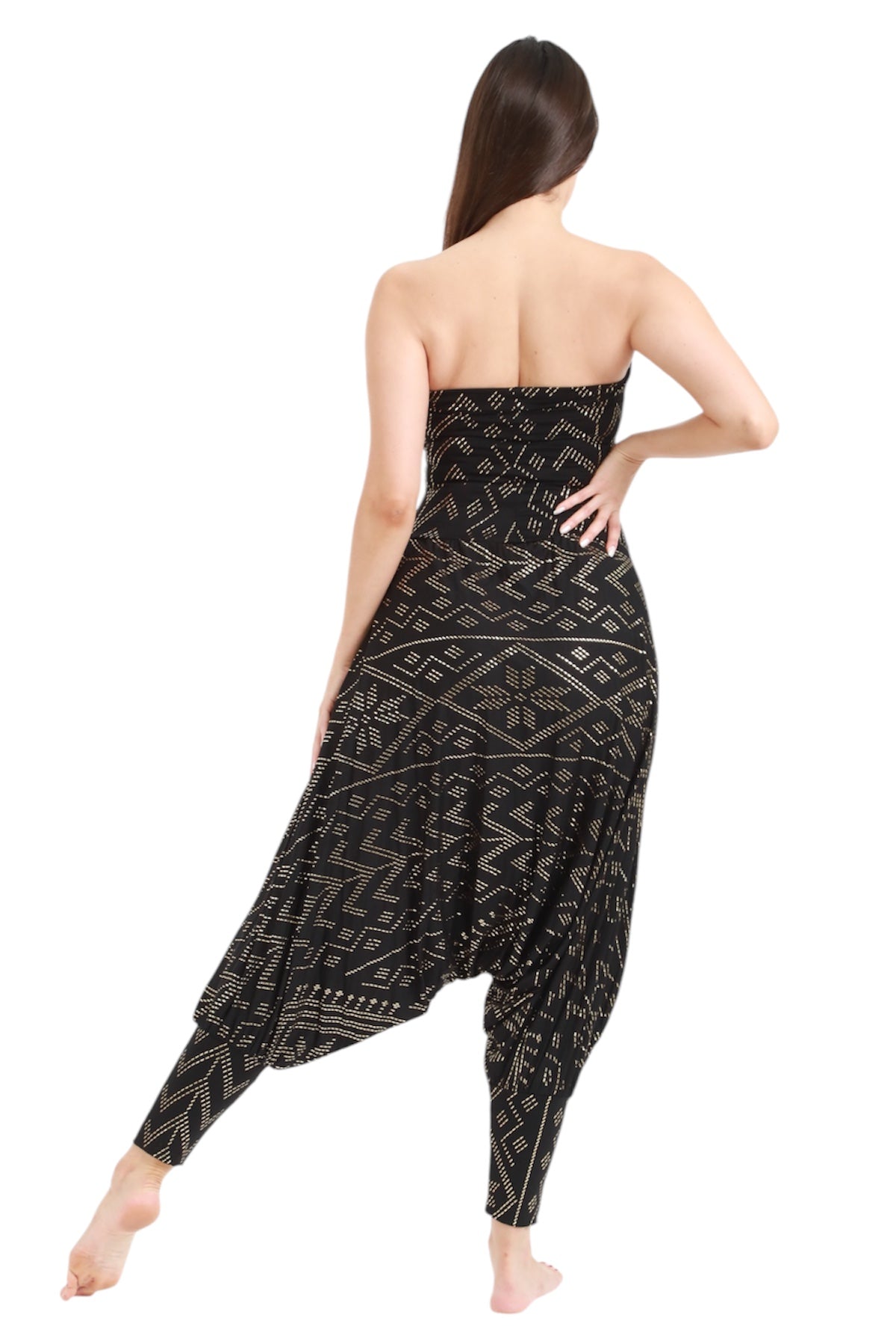 A woman with long brown hair stands barefoot, facing away, wearing the Christine Luxor Harem Jumper by Melodia Designs—a strapless black jumpsuit with beige geometric patterns. One leg is slightly bent and her right hand rests on her hip.