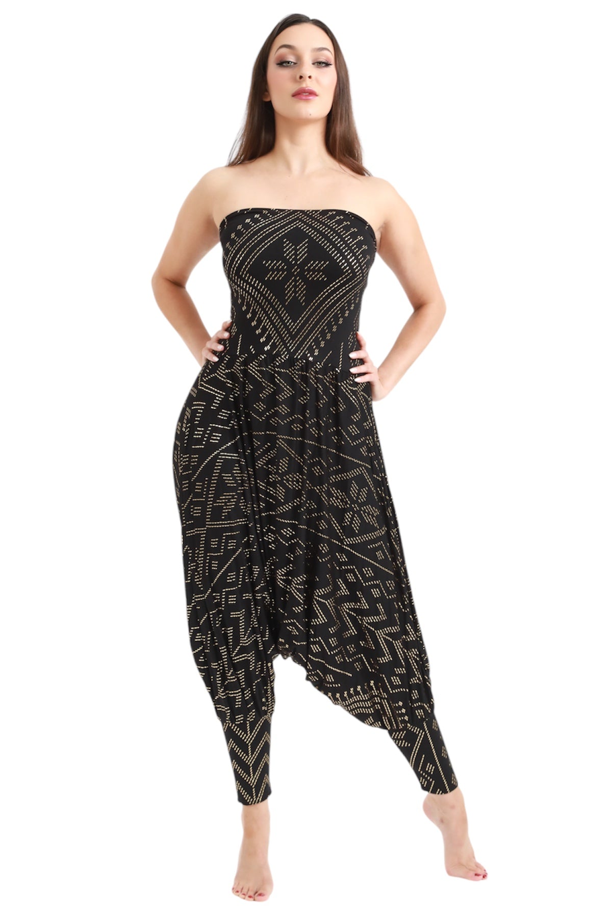 A woman models the Christine Luxor Harem Jumper by Melodia Designs—a strapless, black one-piece with gold geometric patterns and harem pant style—standing barefoot against a white background, hands on hips.
